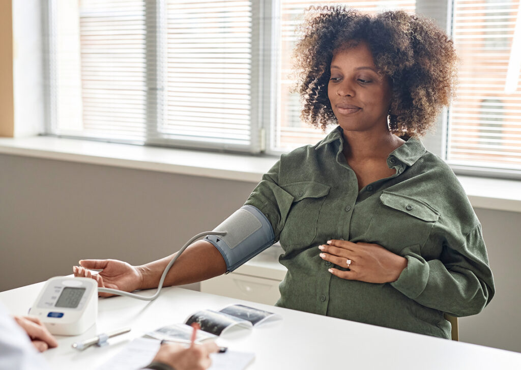 A pregnant woman sitting at a table checking her blood pressure with a digital monitor.