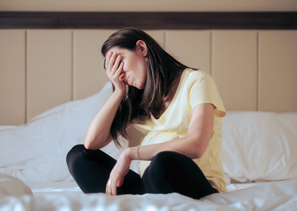 A pregnant woman sitting cross-legged on a bed, holding her hand over her face in distress.