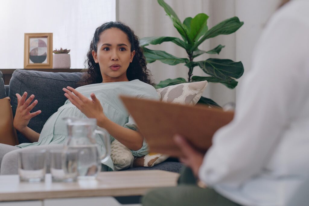 A pregnant woman sitting on a couch gesturing with her hands while talking to a healthcare professional.
