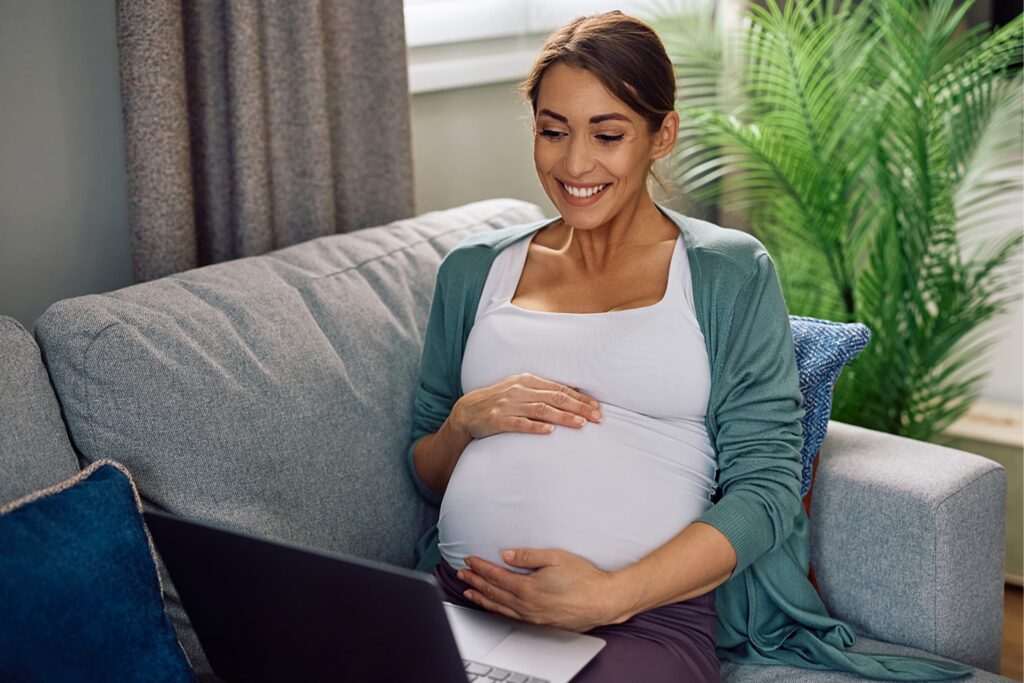 A smiling pregnant woman sitting on a gray couch, gently resting her hands on her stomach while looking down at a laptop screen.