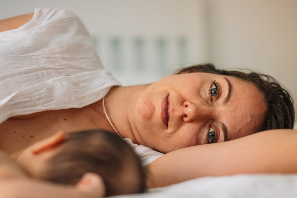 A close-up of a mother lying down in bed, looking tired and sad while resting next to her baby.